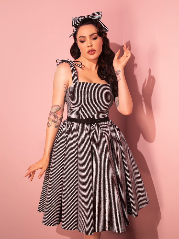 Our stunning female model elegantly displays the Dream House Swing Dress and Matching Hair Bow in Halloween Thin Black and White Stripes, courtesy of Vixen Clothing, the foremost retro dress and vintage clothing brand.
