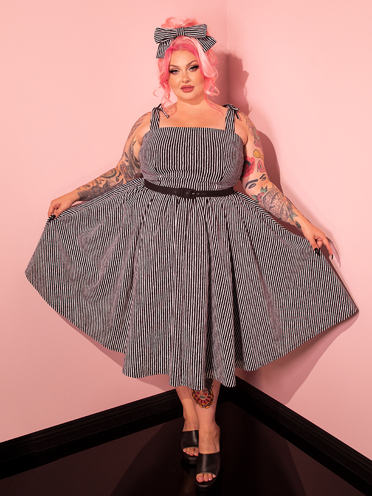 Our beautiful female model exudes elegance while wearing the Dream House Swing Dress and Matching Hair Bow in Halloween Thin Black and White Stripes from Vixen Clothing, the ultimate destination for retro dress and vintage fashion.
