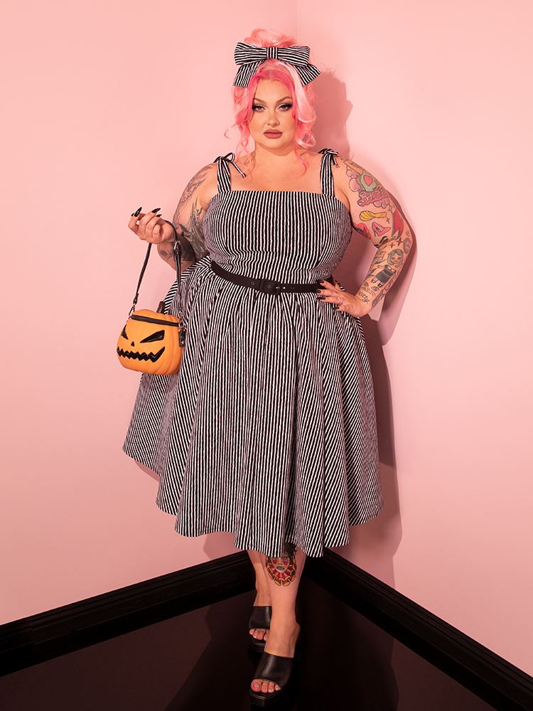 Witness the enchanting beauty of our female model as she dons the Dream House Swing Dress and Matching Hair Bow in Halloween Thin Black and White Stripes, a masterpiece created by Vixen Clothing, the leading retro dress and vintage clothing brand.