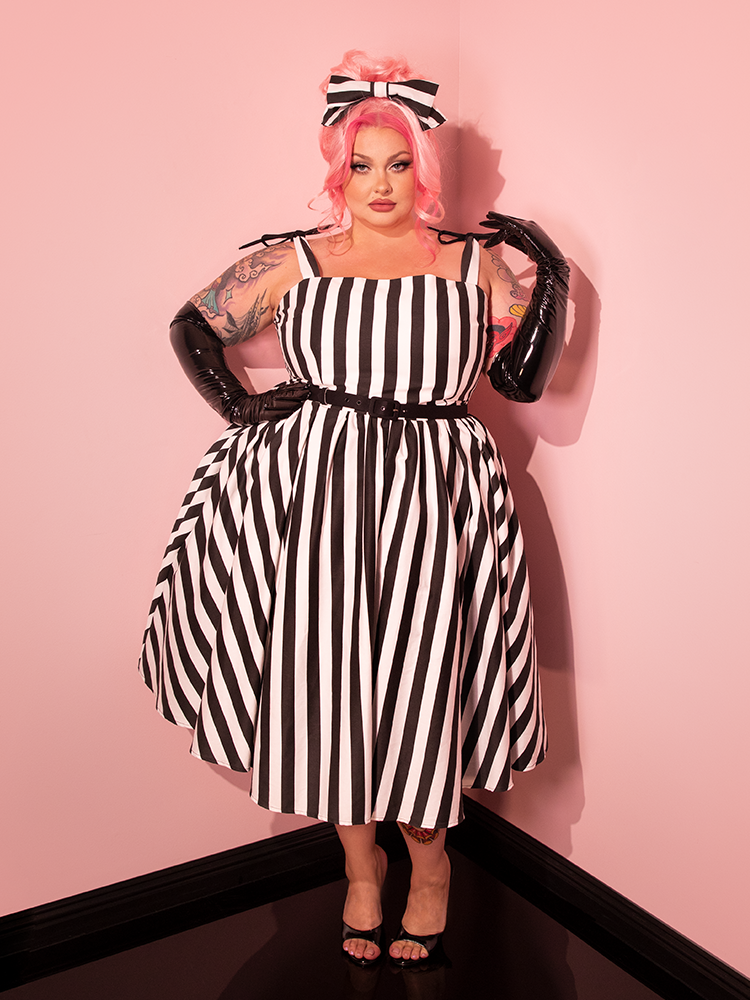 Transport yourself to the retro era with a female model donning the Dream House Swing Dress and Matching Hair Bow in Thick Black and White Stripes, courtesy of the iconic brand Vixen Clothing.