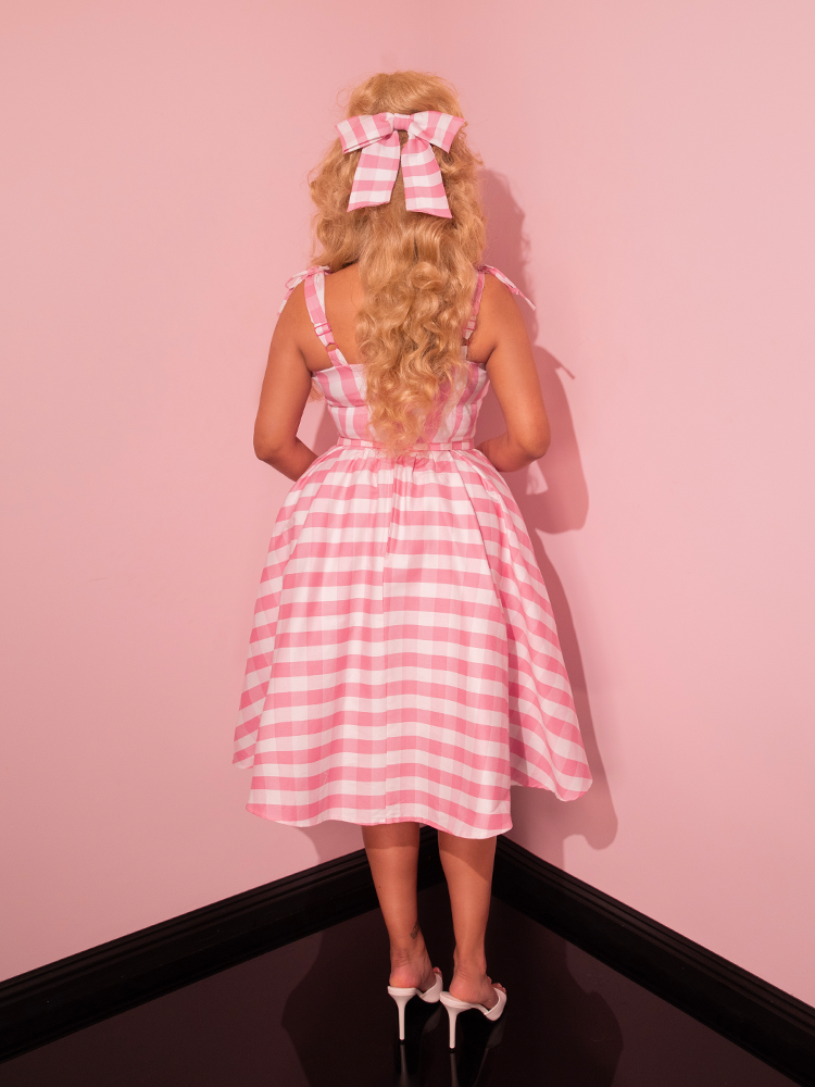 Embark on a retro fashion journey as a female model strikes a pose in an all-pink showroom, donning the Dream-House Swing Dress and Matching Bow in Pink Gingham, a masterpiece by Vixen Clothing, the embodiment of vintage style.