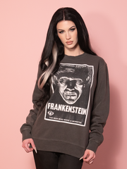A washed black sweatshirt featuring a vintage Frankenstein movie poster graphic, displayed against a pink wall.