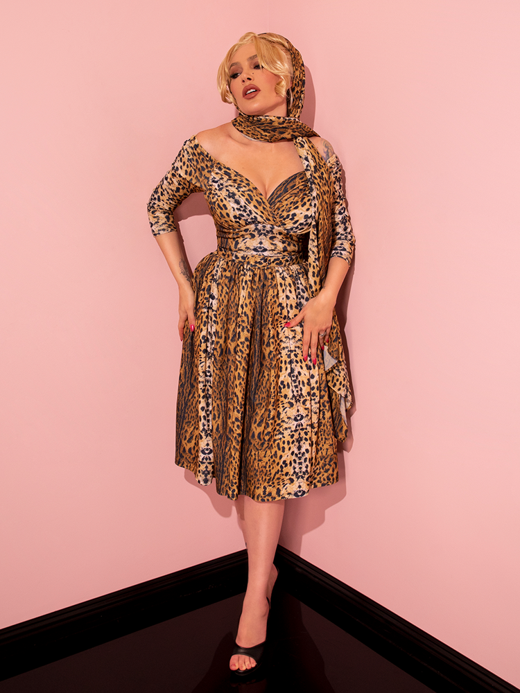Evoking a nostalgic charm, a captivating female model strikes a seductive pose while wearing the Starlet Swing Dress and Scarf in Leopard Print, a gorgeous ensemble from the retro dress brand Vixen Clothing.
