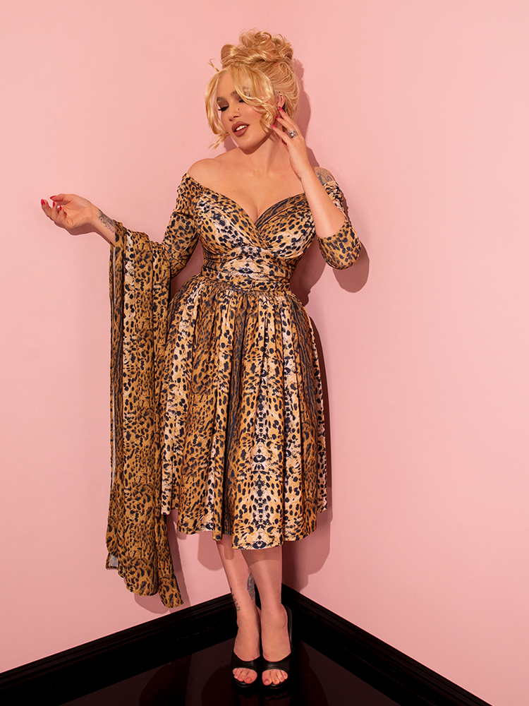 Radiating vintage elegance, a captivating female model strikes a seductive pose while showcasing the Starlet Swing Dress and Scarf in Leopard Print, a timeless creation by Vixen Clothing, a renowned retro dress brand.