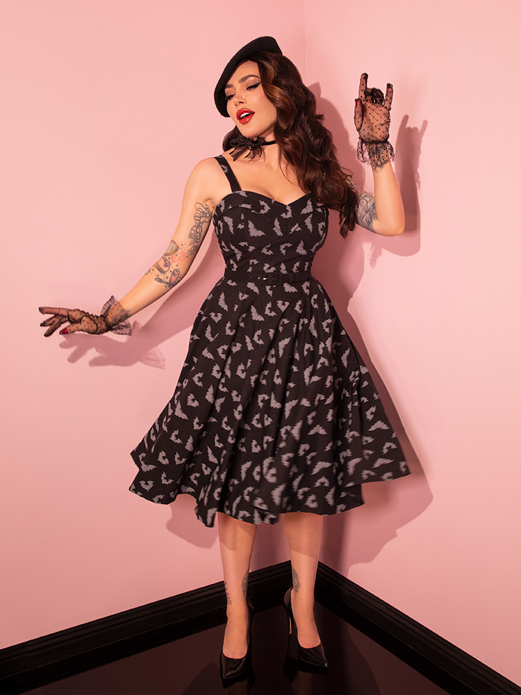 Embrace the charm of retro style as our stunning female model gracefully dons the Maneater Swing Dress, showcasing the Glow in the Dark Bat Print in Black, a creation of Vixen Clothing, the top retro dress brand.