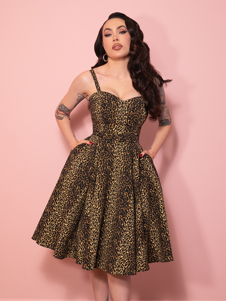 A vintage inspired Maneater Swing Dress in leopard print, styled against a pink wall with timeless glamour.