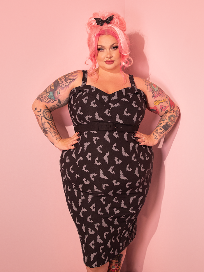 A gorgeously styled model elegantly showcases the Maneater Wiggle Dress in Black Glow in the Dark Bat Print from the retro dress and clothing brand, Vixen Clothing.