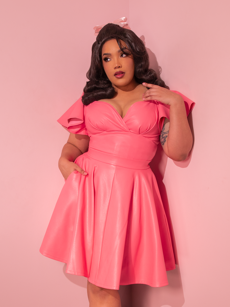 Transporting us to a bygone era, the enchanting model showcases the Flamingo Pink Vegan Leather Bad Girl Skater Skirt, an exquisite vintage creation brought to life by the talented artisans at Vixen Clothing, the ultimate retro dress maker.
