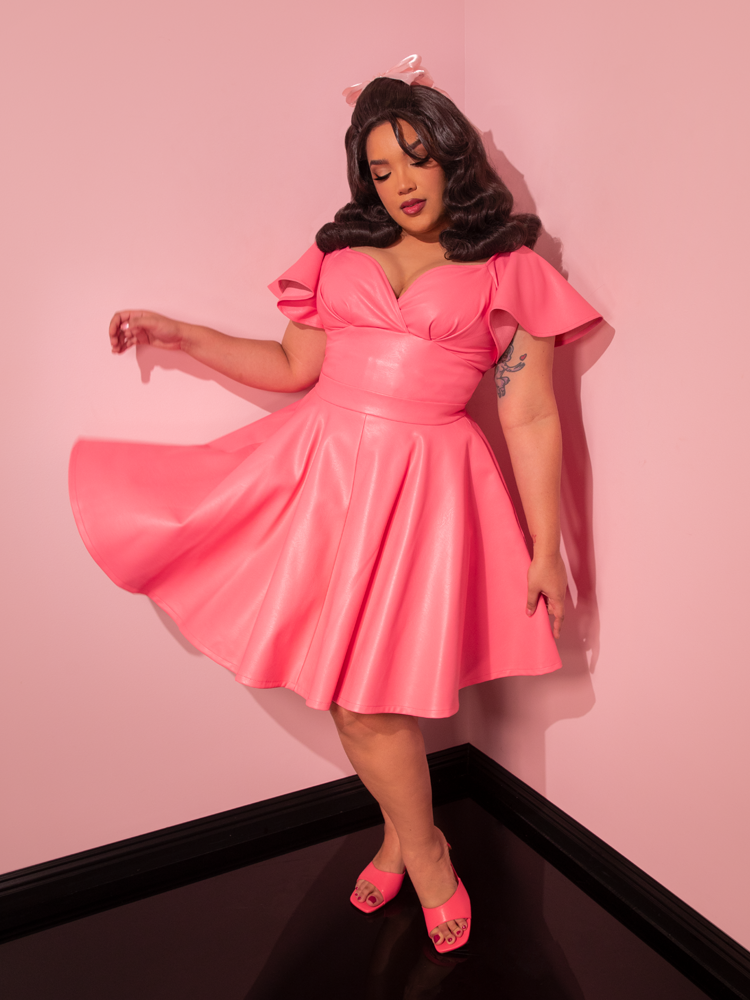 Evoking the spirit of the past, the enchanting model dons the Bad Girl Skater Skirt in Flamingo Pink Vegan Leather, a retro masterpiece that embodies the essence of vintage elegance from Vixen Clothing.
