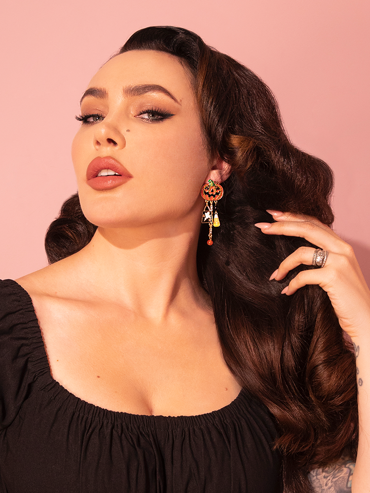 In a stylish moment, the model showcases the all-new Orange Jack O' Lantern Charm Dangle Earrings by Vixen Clothing, the renowned retro fashion label.