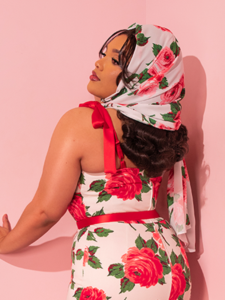 Mesmerizing onlookers, the breathtaking female model effortlessly combines her retro-style dress, accentuated by an enchanting red rose pattern, with the 1950s Inspired Chiffon Rose Scarf from the vintage-style clothing brand Vixen Clothing, epitomizing timeless elegance.
