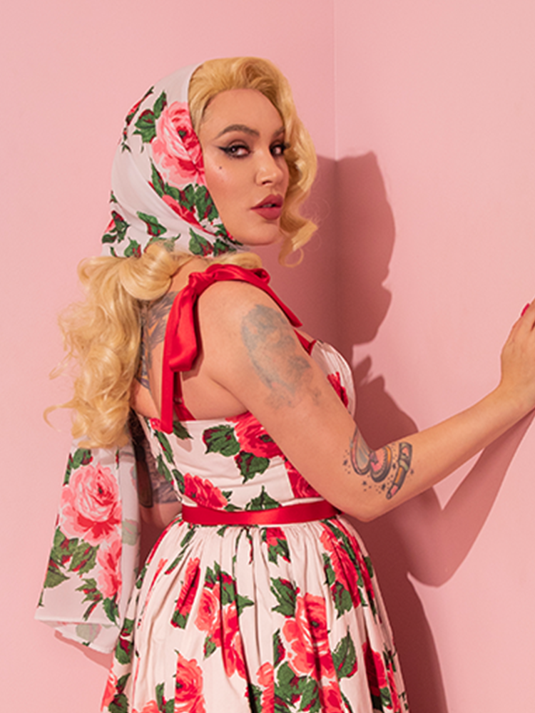 A vision of retro allure, the lovely female model adorns herself in a vintage-inspired dress, exquisitely embellished with a ravishing red rose motif, and expertly complements it with Vixen Clothing's Chiffon Rose Scarf, evoking the essence of the 1950s fashion revival.