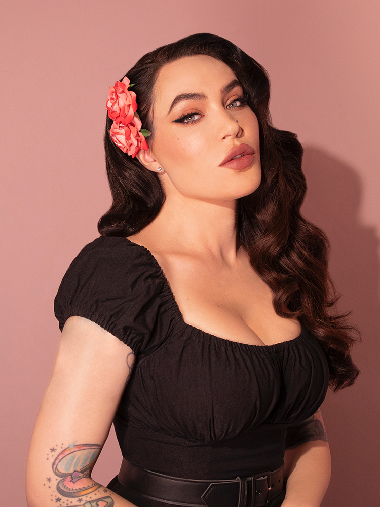 Micheline Pitt gazes into the camera while wearing the Vintage-Style Double Rose Hair Comb in Pink/Red from vintage style clothing brand Vixen Clothing.