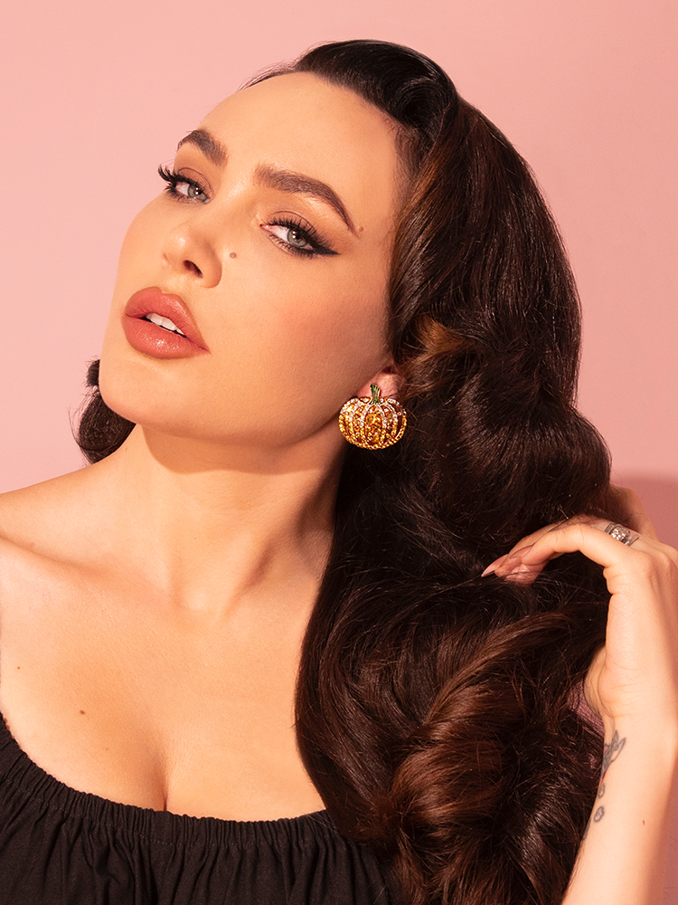 Radiant in her elegance, a female model showcases the Rhinestone Jack-O' Lantern Earrings in Gold, a creation of the retro fashion brand Vixen Clothing.