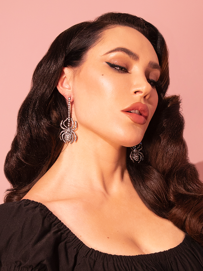 Micheline Pitt gracefully showcases Vixen Clothing's Silver Rhinestone Spider Dangle Earrings, adding a touch of vintage charm to her ensemble.