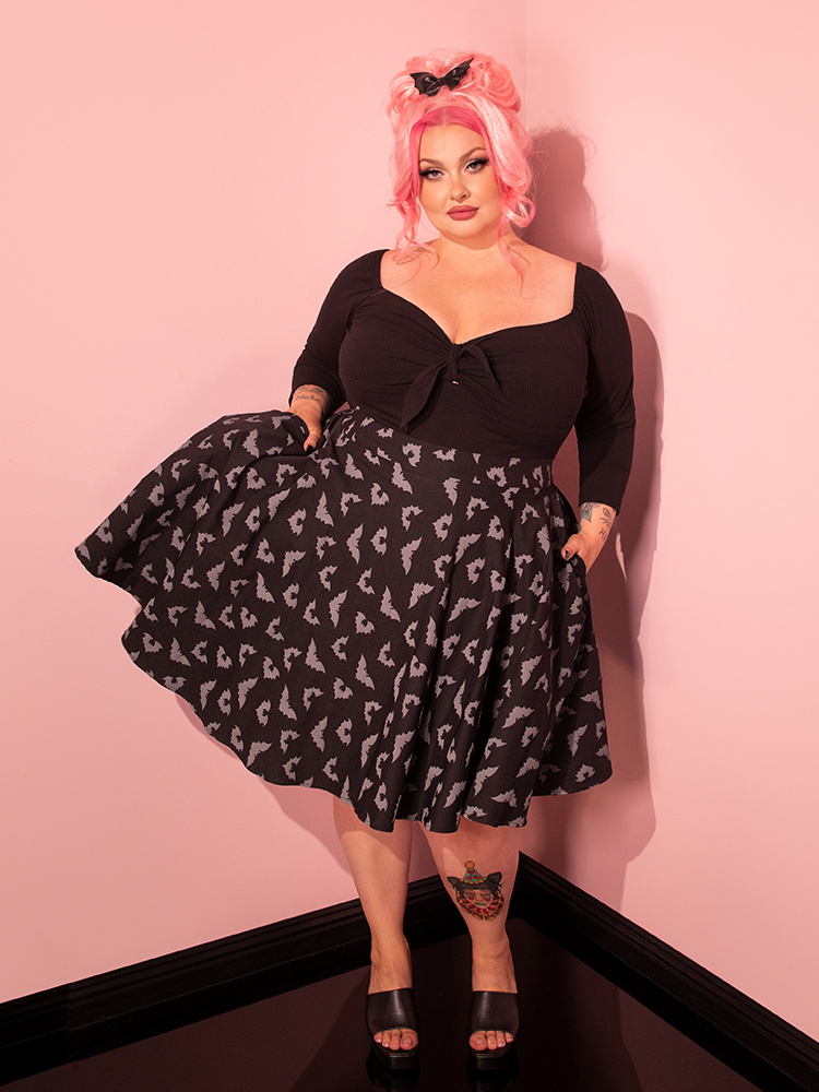 A vintage-inspired pin-up sensation struts her stuff, wearing the Maneater Skater Skirt in its enchanting Glow-in-the-Dark Bat Print, amidst the nostalgic ambiance of an all-pink showroom.