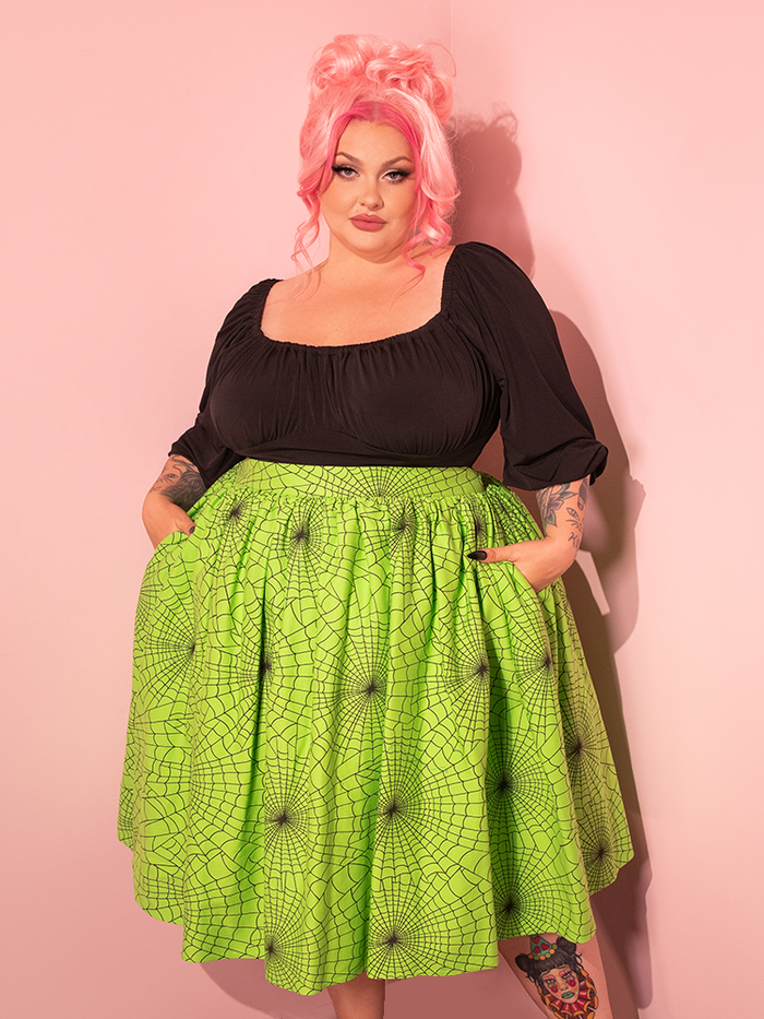 Radiating vintage charm, a female model elegantly displays the Vixen Swing Skirt in Slime Green Spider Web Print, courtesy of the retro fashion label Vixen Clothing.