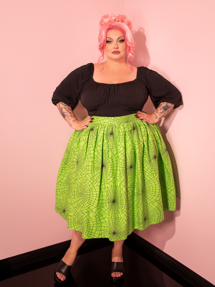 With captivating allure, a female model highlights the Vixen Swing Skirt in Slime Green Spider Web Print, a creation of the renowned retro clothing brand Vixen Clothing.