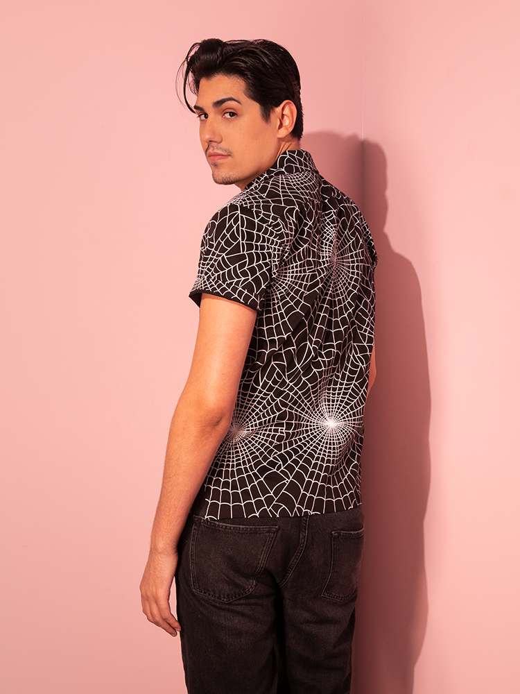 In a striking display, a male model wears the Button Up Short Sleeve Shirt in Halloween Spider Web Print (Black) from the retro-style brand Vixen Clothing.