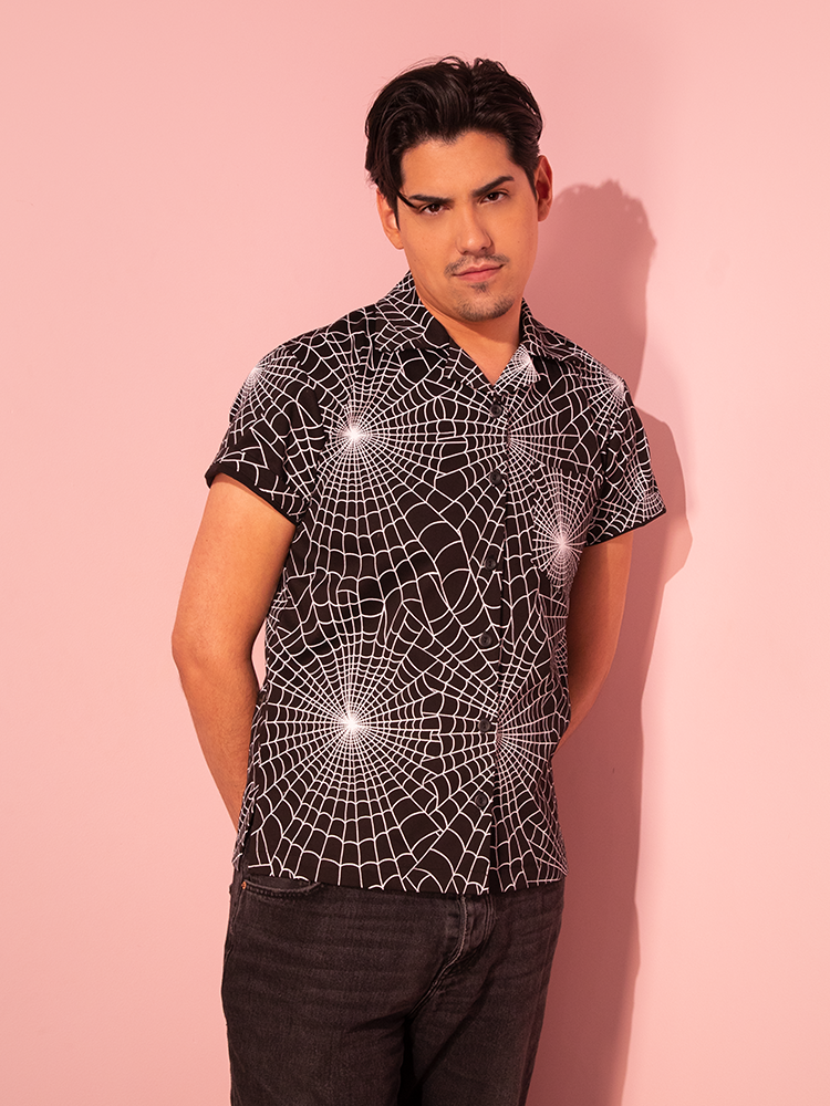 A male model showcases the Halloween Spider Web Print Button Up Short Sleeve Shirt in Black from the vintage-inspired fashion label Vixen Clothing.