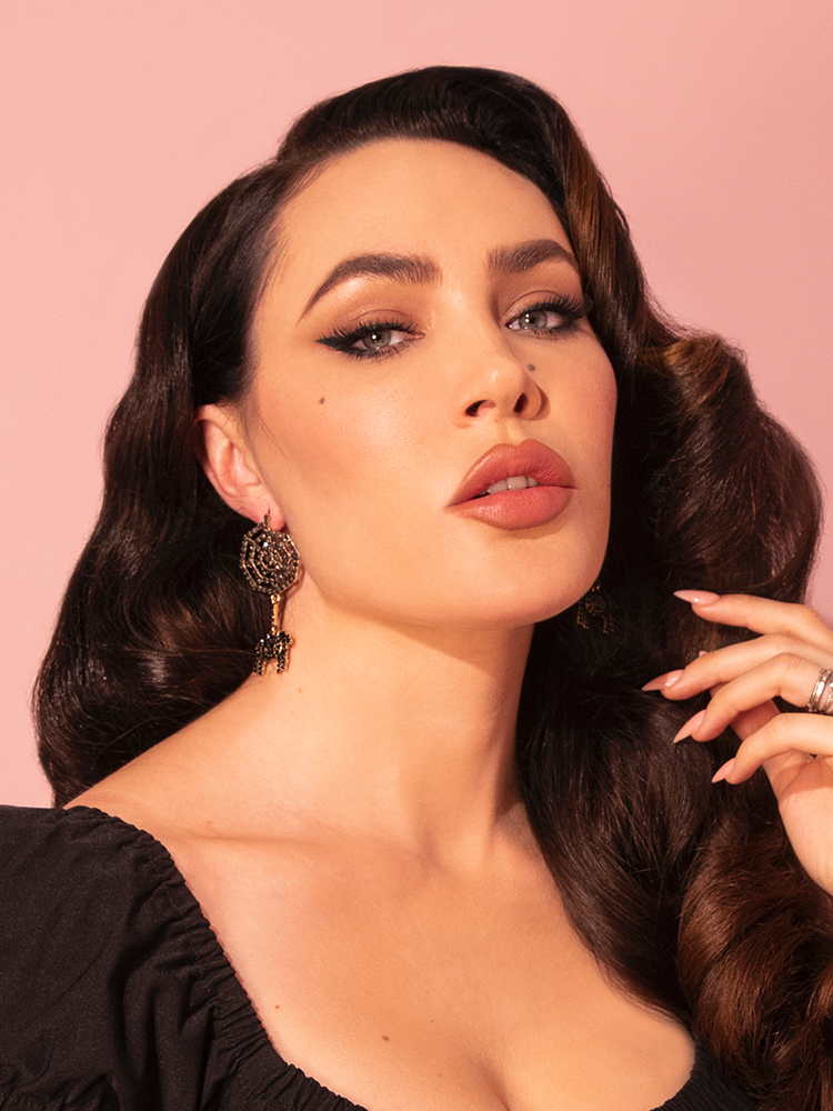 Micheline Pitt stares into the camera while pulling back her hair on one side to show the Rhinestone Spider and Web Dangle Earrings in Black from vintage clothing brand Vixen Clothing.
