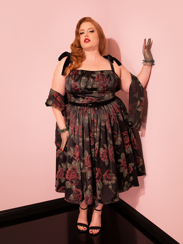 With grace and poise, the retro model presents the 1950s Satin Swing Sundress and Scarf in Black Vintage Roses from Vixen Clothing, embodying its timeless charm.