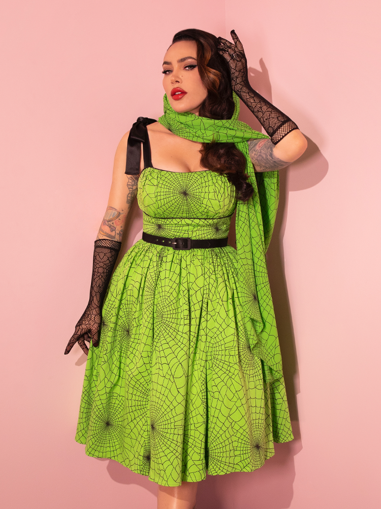 Immerse yourself in the world of style with a gorgeous female model elegantly wearing the 1950s Swing Sundress and Scarf in Striking Slime Green Spider Web Print, a creation of Vixen Clothing's retro flair.
