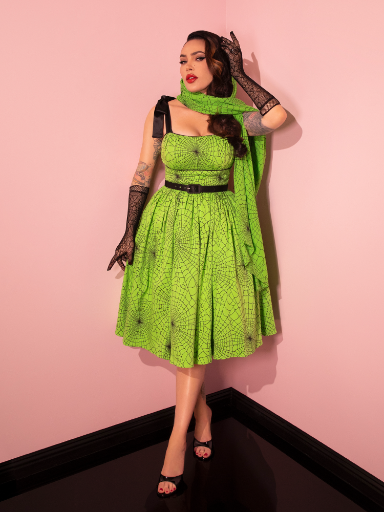 Embark on a journey through vintage aesthetics as a beautiful female model dons the 1950s Swing Sundress and Scarf, featuring Playful Slime Green Spider Web Print by the iconic retro dress and clothing brand, Vixen Clothing.