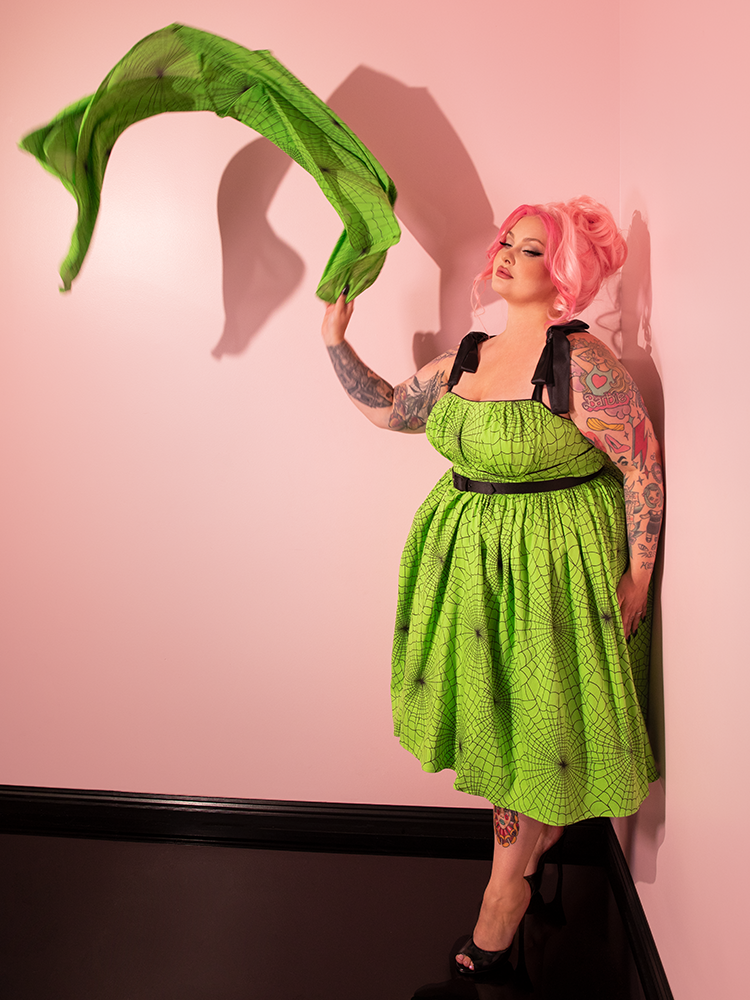 Experience the glamour of the past with a beautiful female model showcasing the 1950s Swing Sundress and Scarf in Bold Slime Green Spider Web Print, crafted by Vixen Clothing's retro expertise.
