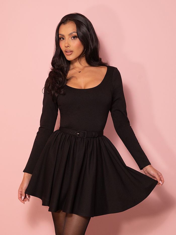 A vintage-inspired black swing mini dress displayed against a pink wall, highlighting its flared skirt and elegant neckline that echo classic Hollywood glamour.