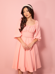A vintage style dress in ballet pink shown against a pink wall, highlighting its soft elegance and timeless charm.