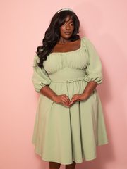 A vintage inspired vacation dress in matcha green flows gracefully against a pink wall, capturing breezy elegance.