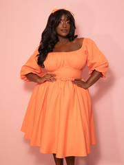 A vintage inspired vacation dress in sherbet orange glows brightly against a pink wall, capturing playful vintage elegance.