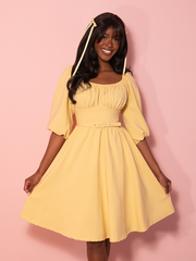 A vintage inspired dress in soft yellow with a row of covered buttons and puffed sleeves exudes classic Hollywood charm against a pink backdrop.