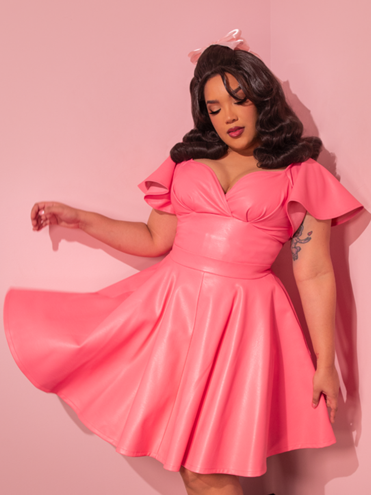 With a touch of nostalgia, the gorgeous model mesmerizes in the Flamingo Pink Vegan Leather Bad Girl Skater Skirt, an enchanting vintage-inspired garment meticulously crafted by the renowned retro dress maker Vixen Clothing.