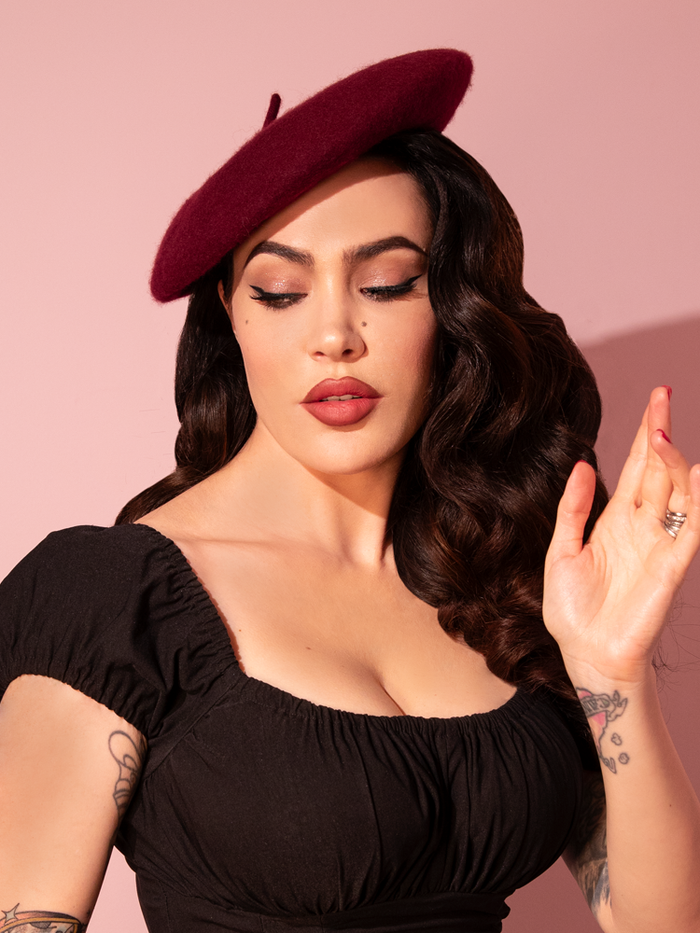 Micheline Pitt posing playfully with one hand raised, wears the Vintage-Style Beret in Burgundy Wine paired with a low-cut black retro top from Vixen Clothing.