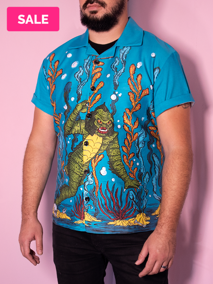 Male model standing against pink background wearing a teal blue button-up shirt with a print of an underwater scene with the swamp creature.