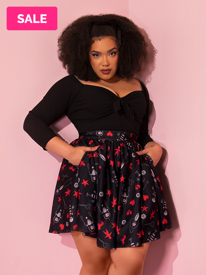 Ashleeta tucks her hands into the EDWARD SCISSORHANDS Skater Skirt in “I am not complete” Novelty Print while wearing a black low-cut top tucked in.