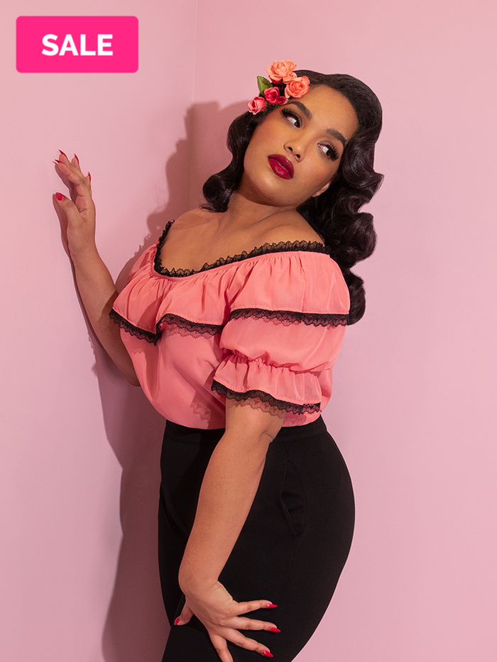 A closeup of Ashleeta looking over her shoulder wearing pink flowers in her hair modeling the Vixen chiffon peasant top in peach with black lace.