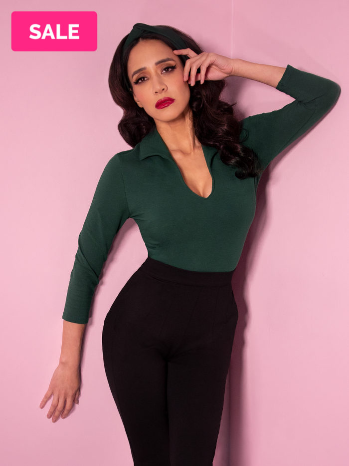 Milynn, with her hand on her face, modeling the Vixen top in hunter green paired with black vintage style pants.