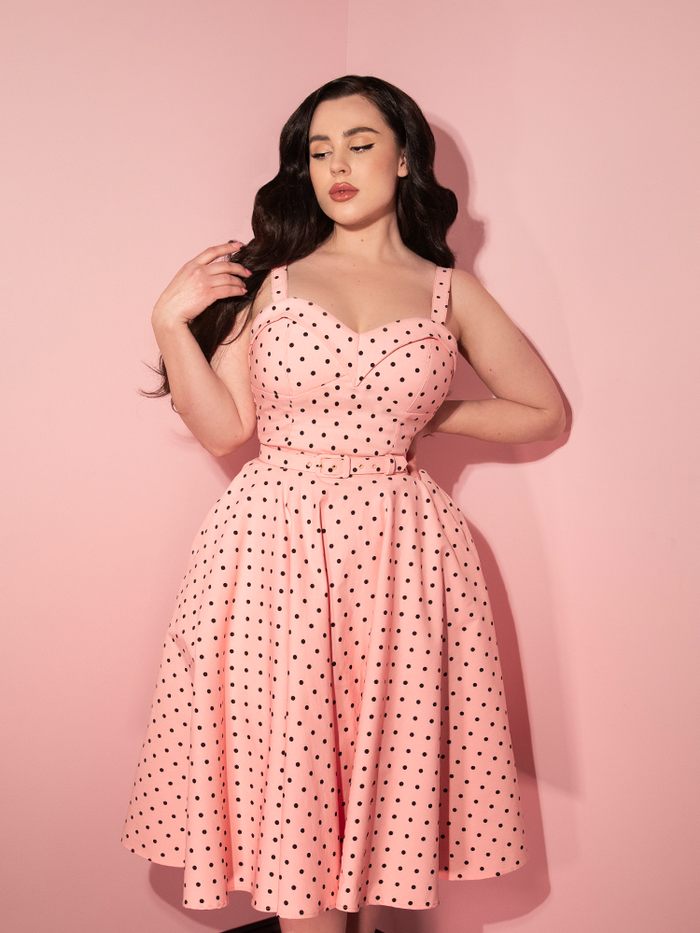 Rachel Sedory in the Maneater Swing Dress in Rose Pink Polka Dot while looking away and twirling her hair.