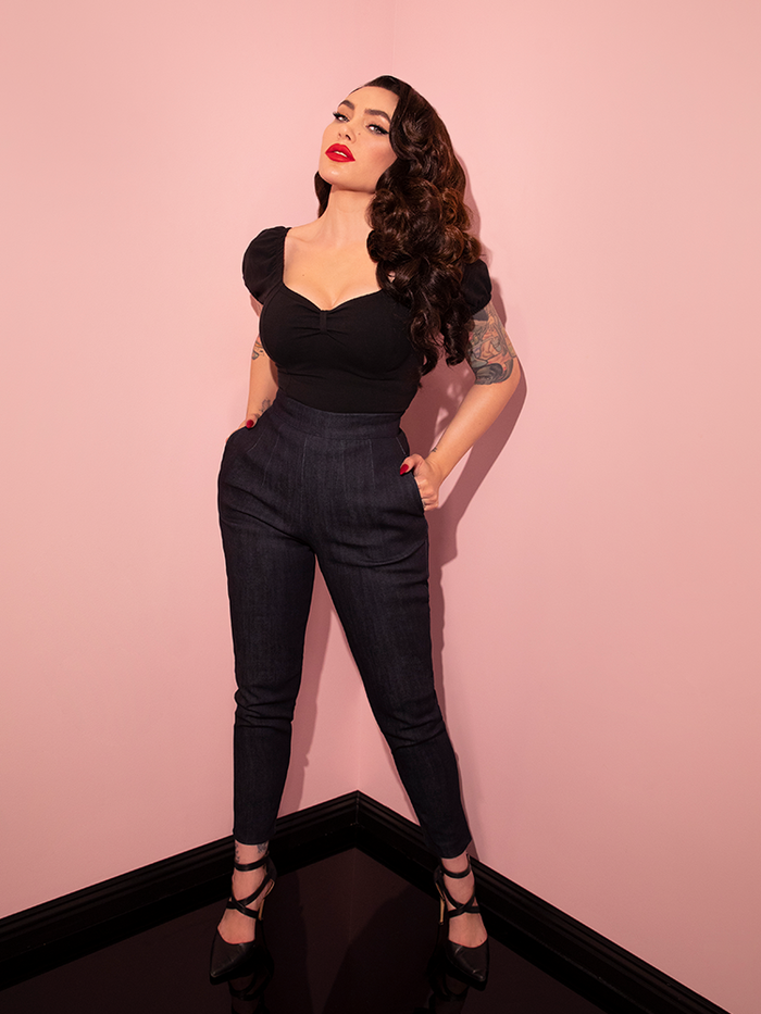 Full length shot of Micheline Pitt standing in the corner of her showroom tucking her hands into the side pockets of the Cigarette Pants in Denim that she has paired with a low-cut black top.
