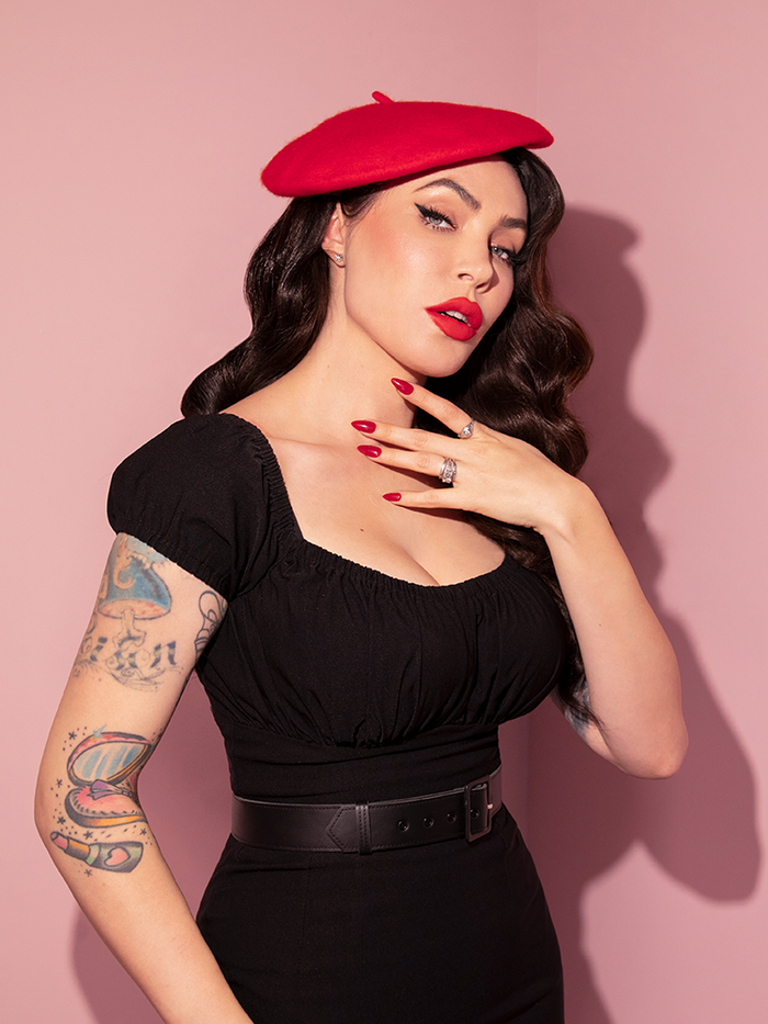 Waist level shot of Micheline Pitt wearing the Vintage Style Beret in Classic Red Media with a black low-cut top and black pants.