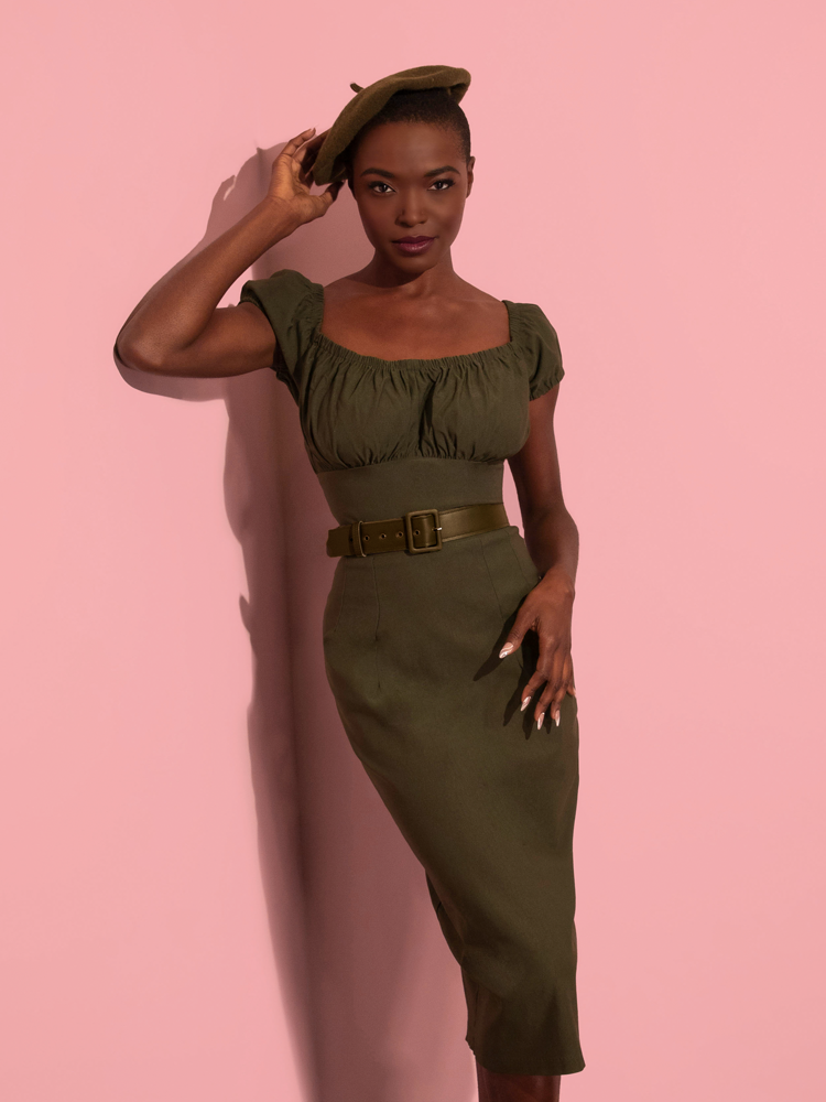 Model wearing the Peasant Wiggle Dress in Olive Green with matching beret standing in all-pink room.