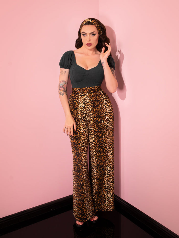 Full length shot of Micheline Pitt standing in the corner of her pink showroom wearing the Vacation Pants in Leopard Crepe with a low-cut retro top tucked in.