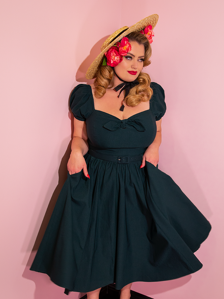 A closeup of Blondie wearing a straw hat and flowers in her hair modeling the Vixen dress in hunter green with her hands in her pockets.