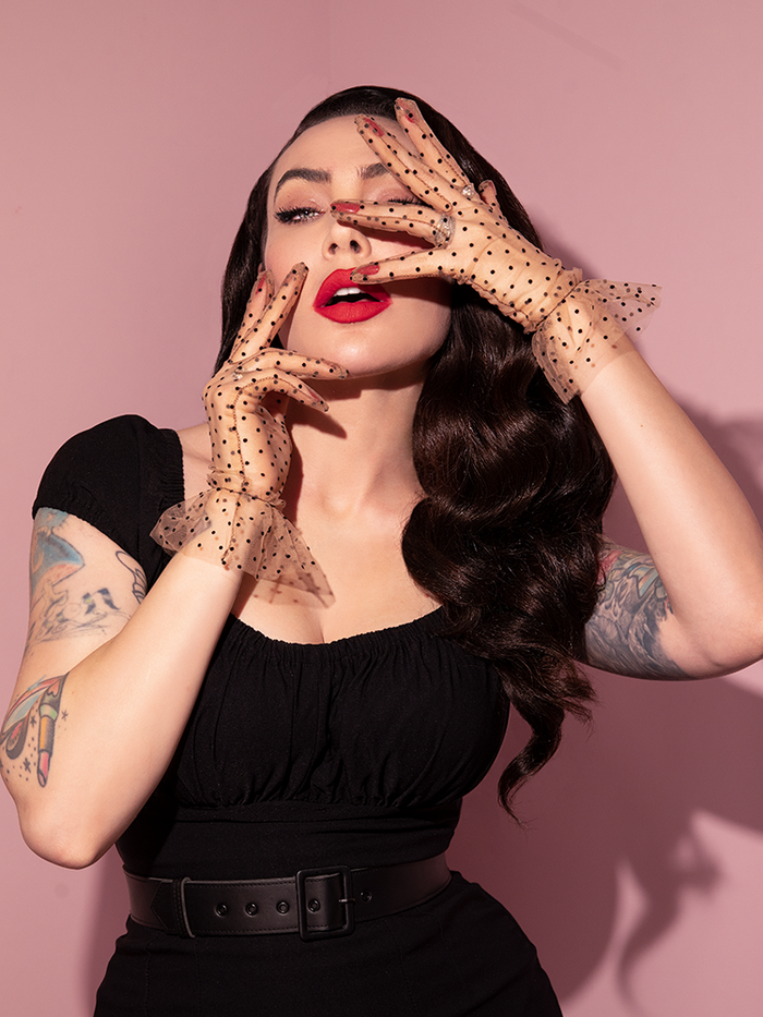 Micheline Pitt seductively poses her hands across her face while wearing the Mesh Polka Dot Gloves in Tan from retro clothing brand Vixen Clothing.