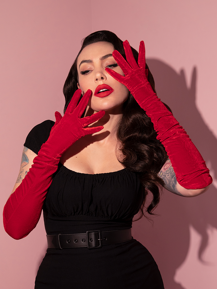 Micheline Pitt strikes a dramatic pose with her hands slightly obscuring her face while donning the Velvet Gloves in Red from retro clothing company Vixen Clothing.