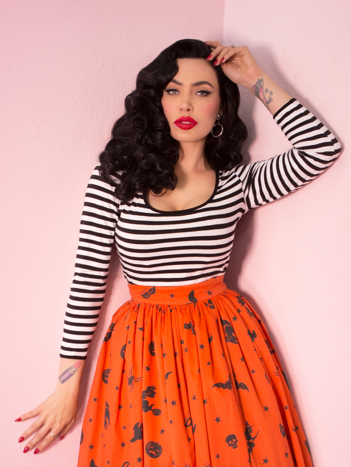 Waist level shot of Micheline Pitt in the Troublemaker Top in Black and Whites Stripes paired with a pumpkin orange Halloween skirt. 