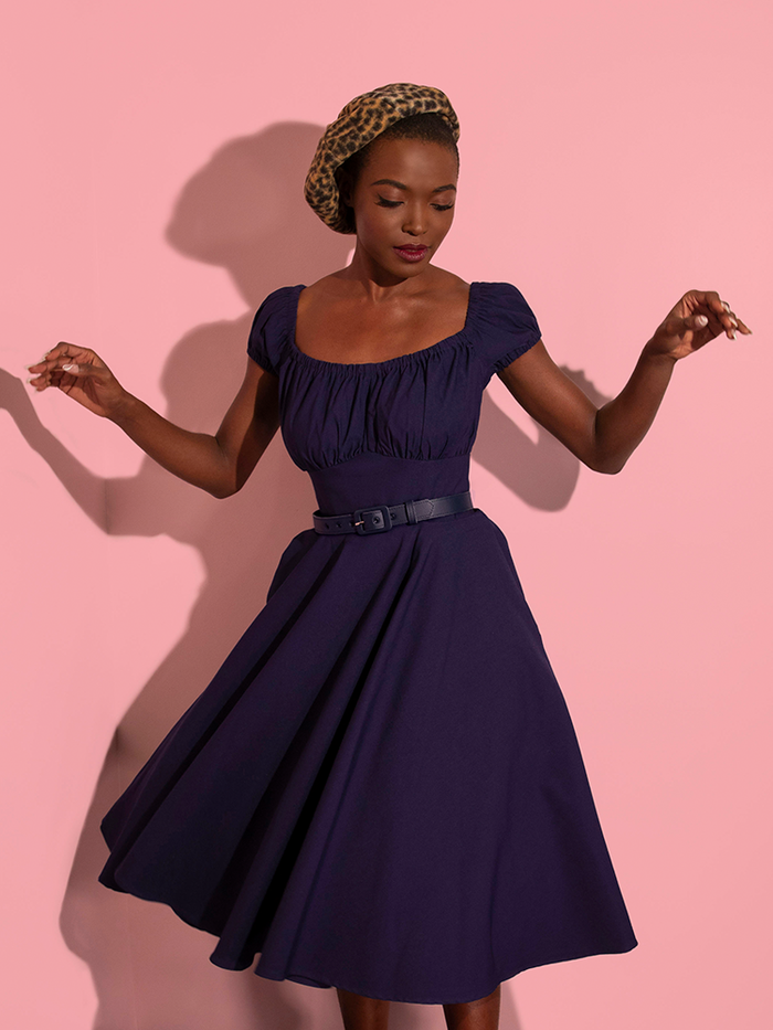 Model is photographed twirling in the Peasant Swing Dress in Navy while also wearing a leopard print beret.